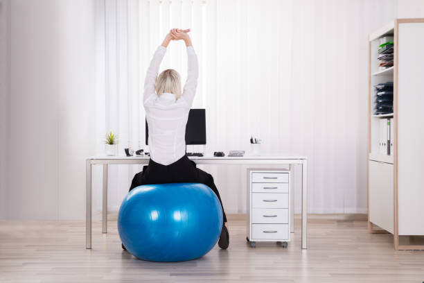 Rear View Of Businesswoman Stretching Her Arms Rear View Of Businesswoman Sitting On Fitness Ball Stretching Her Arms In Office yoga ball work stock pictures, royalty-free photos & images