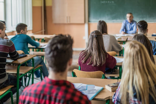 Rear view of high school students attending a class. Back view of large group of students sitting in the classroom and listening to their teacher. Focus is on girl in purple sweater. high schools stock pictures, royalty-free photos & images