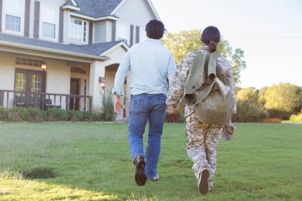 Rear view of husband leading military wife back home In this full length rear view, a husband and wife hold hands as they walk across their front lawn to their home.  The wife wears a military camouflage uniform and carries a MOLLE. veterans returning home stock pictures, royalty-free photos & images
