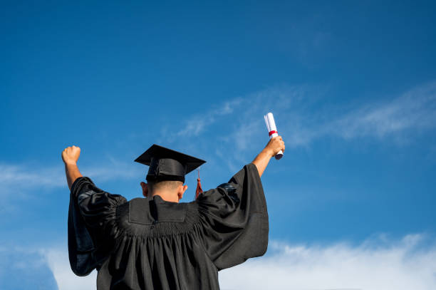 Rear View Of man Throwing hands up a certificate and Cap in the air, Graduation day on sky background stock photo