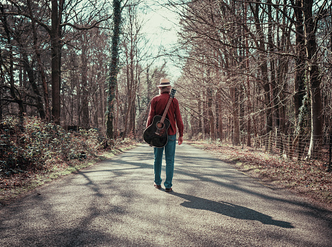 Color image depicting the rear view of a man wearing a red check shirt, denim jeans and a straw hat while walking on a beautiful tree lined country road. He is enjoying the freedom of exploring and wandering in nature, and he has an acoustic guitar strapped to his back. Room for copy space.
