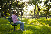 Rear view of one mature woman sitting on a park bench in the summer season.  Bright sunny day with lush green grass and trees.  The woman gazes off into the distance as she relaxes on a beautiful day.  Solitude, lonliness, contemplation.  She has short blond hair and wears a purple shirt and jeans.   Copyspace to right in this tranquil nature scene.