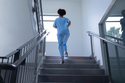 Rear view of mixed race female doctor wearing scrubs running up stairs in hospital. medical professional at work.