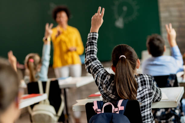 Rear view of schoolgirl raising her arm to answer the question in the classroom. Back view of elementary student raising arm in order to answer a questing during a class. elementary age stock pictures, royalty-free photos & images