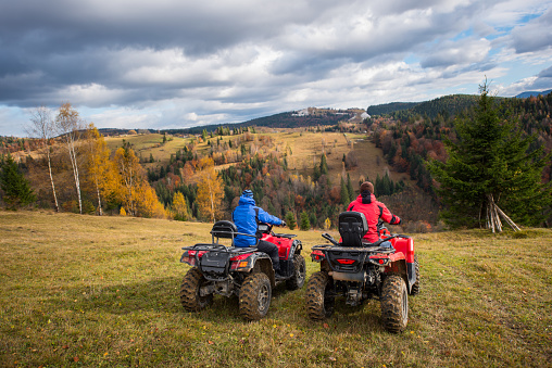 Rear view of two men sitting on quad bikes enjoying beautiful landscape of mountains and colorful forest under the sky with cumulus clouds in autumn