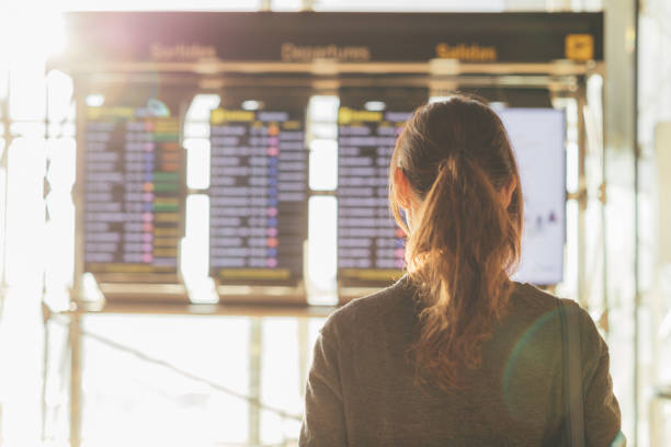 Rear view of young woman looking at flight information board in airport stock photo