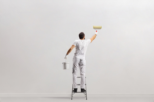 Rear view shot of a painter holding a bucket and painting a wall on a leader isolated on white background