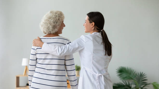 Rear view smiling girl caregiver supporting older woman, touching shoulders Rear view smiling girl caregiver supporting older woman, touching shoulders, hugging, expressing care and love, good relations, nurse wearing white uniform coat comforting mature patient, healthcare patience stock pictures, royalty-free photos & images