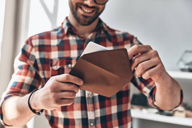 Receiving greeting card. Close up of young man opening envelope and smiling while standing indoors sending stock pictures, royalty-free photos & images