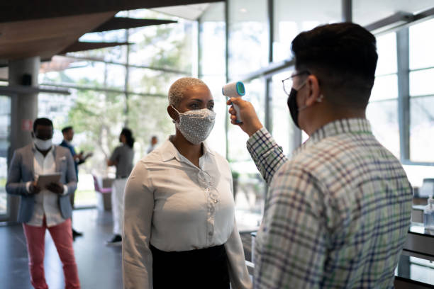 Receptionist measuring temperature of female employee at office's entrance - with face mask Receptionist measuring temperature of female employee at office's entrance - with face mask returning stock pictures, royalty-free photos & images