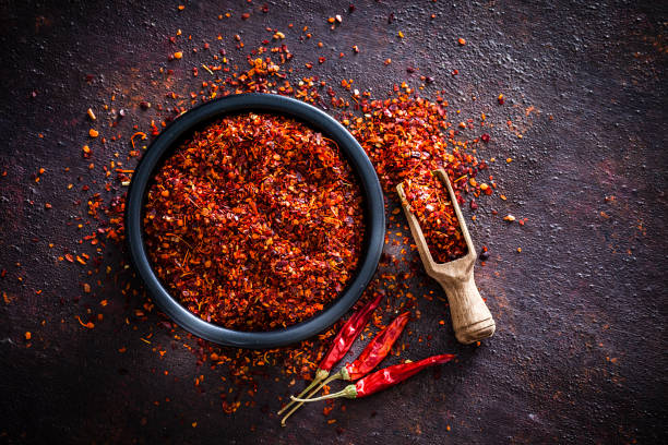 Red chili pepper flakes shot from above Spices: Top view of a black bowl filled with red chili pepper flakes shot on abstract brown rustic table. A wooden serving scoop with pepper flakes is beside the bowl, pepper flakes are scattered on the table. Three dried red chili peppers are beside the bowl. Predominant colors are brown and red. Low key DSRL studio photo taken with Canon EOS 5D Mk II and Canon EF 100mm f/2.8L Macro IS USM. chili pepper stock pictures, royalty-free photos & images