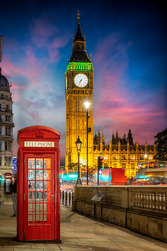 Red telephone booth in front of the illuminated Big Ben clocktower in London, United Kingdom, just after sunset with street traffic