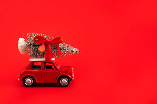 Red toy car carrying Christmas tree with red ribbon on top of the roof on red background. Space for text on the right stock photo