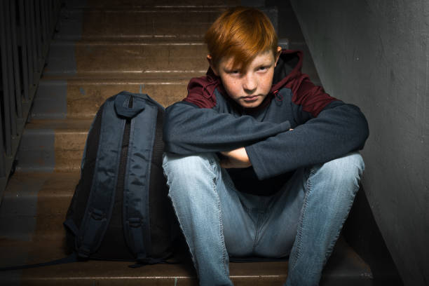 A red-haired boy, a teenager of twelve, with an anxious expression on his face, sits on the ladders at the entrance of a residential building. Children's fear and anxiety, family social problems. stock photo