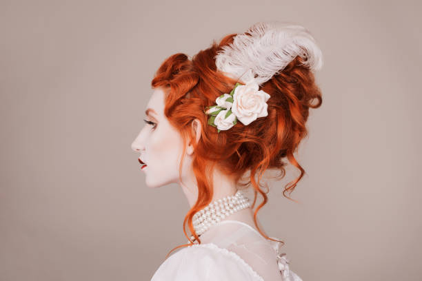 Red-haired woman in white dress with pale skin on gray background.  A vampire woman with a beautiful hairdo with a feather and flowers in her hair  victorian gown stock pictures, royalty-free photos & images