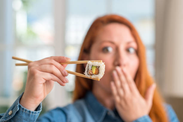 Redhead woman eating sushi using chopsticks cover mouth with hand shocked with shame for mistake, expression of fear, scared in silence, secret concept Redhead woman eating sushi using chopsticks cover mouth with hand shocked with shame for mistake, expression of fear, scared in silence, secret concept how do you say shut up in japanese stock pictures, royalty-free photos & images