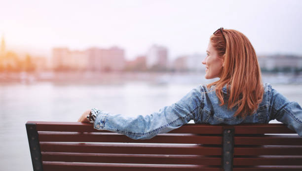 Redhead woman relaxing on bench at riverside in orange sunlight stock photo