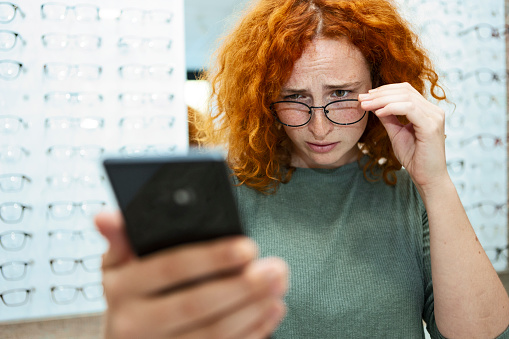 Redhead young woman with eyesight problems trying to read phone text