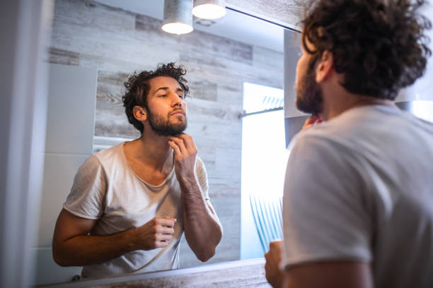 réflexion de l'homme beau avec la barbe regardant le miroir et le visage touchant dans le toilettage de salle de bains - homme miroir photos et images de collection
