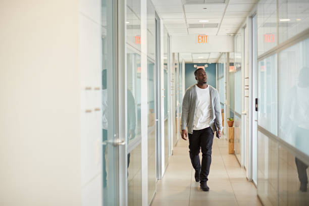 Relaxed Black Freelancer Walking in Miami Coworking Office Full length front view of man in mid 20s wearing casual clothing and approaching camera while walking in office hallway carrying laptop. approaching stock pictures, royalty-free photos & images
