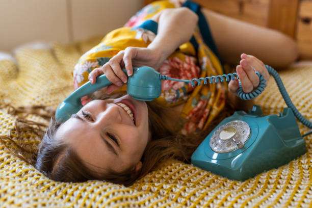 Relaxed cute blond young woman in bed late in the morning using a vintage phone to talk to a friend stock photo
