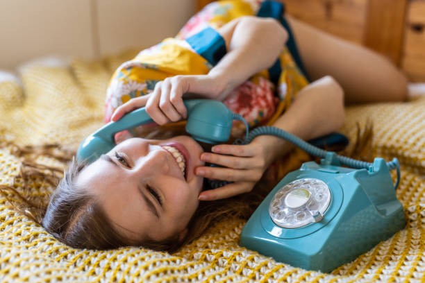 Relaxed cute blond young woman in bed late in the morning using a vintage phone to talk to a friend stock photo