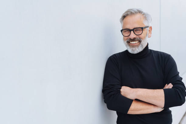 Relaxed self-assured senior man with beard Relaxed self-assured senior man with beard and glasses standing with folded arms in front of a receding white wall mid adult stock pictures, royalty-free photos & images