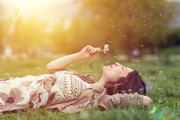 Relaxed woman in the park blowing dandelion Young woman lying down in the grass and blowing dandelion lying down stock pictures, royalty-free photos & images