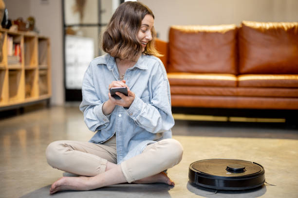 Relaxed woman with phone and robotic vacuum cleaner stock photo