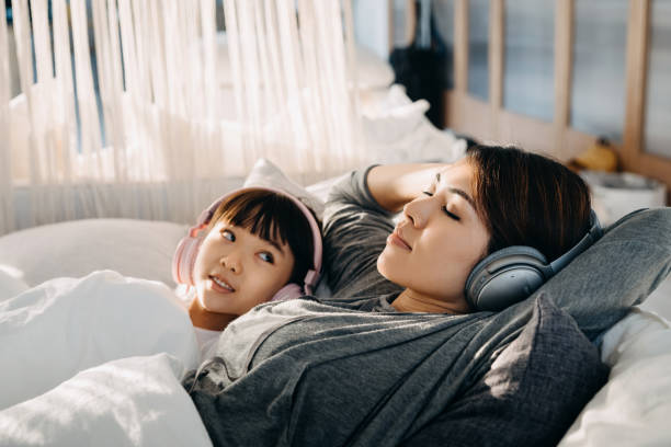 Relaxed young Asian mother and little daughter lying on the bed listening to music with headphones while cheeky little daughter peeking at her mother lazy mom stock pictures, royalty-free photos & images