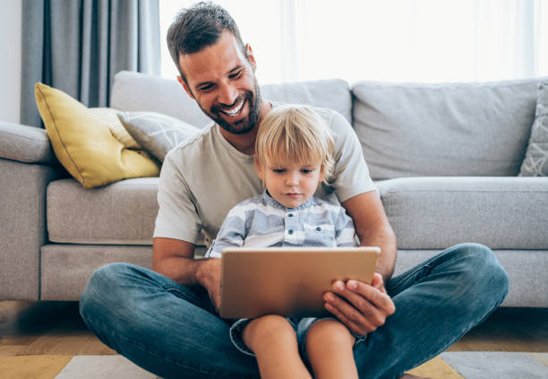 Relaxing at home with wireless technology. Shot of a smiling young father and his cute little son using digital tablet while sitting on the floor at home. Happy father and son making video call with tablet at home. fathers day stock pictures, royalty-free photos & images
