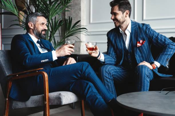 Relaxing. Full length of two young handsome men in suits holding glasses and looking at each other while resting indoors. Relaxing. Full length of two young handsome men in suits holding glasses and looking at each other while resting indoors high society stock pictures, royalty-free photos & images