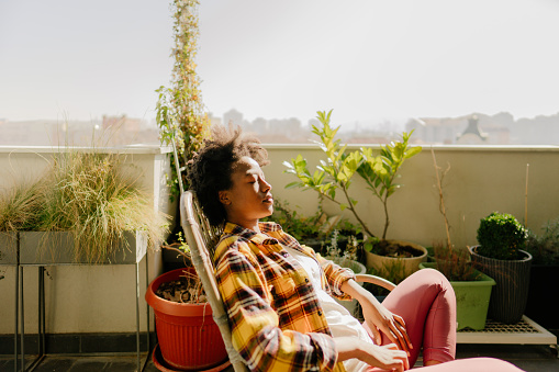 Photo of a young woman sitting and enjoying the tranquility of her lovely rooftop urban garden, on a bright spring morning.