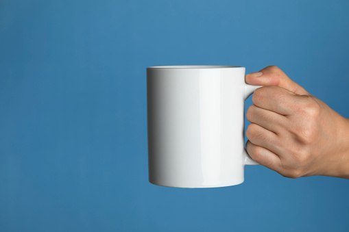Caucasian female is holding a white coffee mug in hand in front of a blue background.