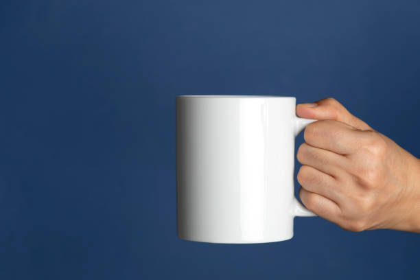 Relaxing Caucasian female is holding a white coffee mug in hand in front of a navy blue background. mug stock pictures, royalty-free photos & images