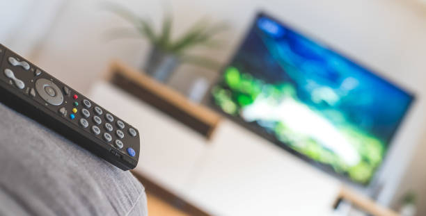 Remote control on the sofa, streaming on a smart TV. TV remote control in the foreground, tv in the blurry background. Streaming. video on demand stock pictures, royalty-free photos & images