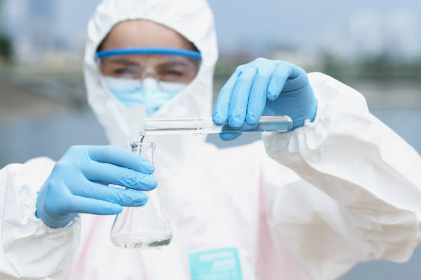 Researcher in protective suit and glasses with gloves pours water from test tube glass beaker stock photo