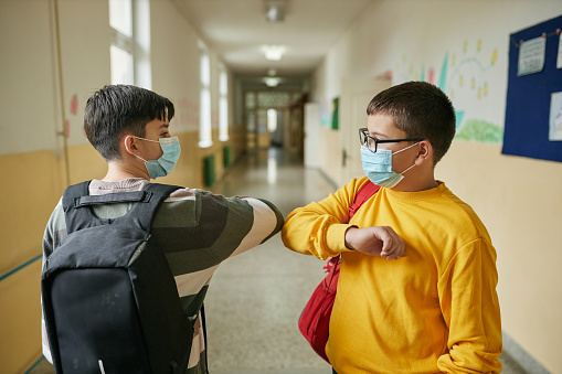 Cheerful elementary students waiting in a school hall before class starts while wearing protective face mask during COVID-19 pandemic