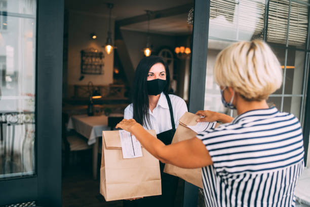 Restaurant employer giving packed food away to a customer. Reopening after COVID-19 quarantine concepts. A kind waiter wearing protective face mask is giving a disposable pack with food to a female customer. Reopening after COVID-19 quarantine concepts. take out food stock pictures, royalty-free photos & images