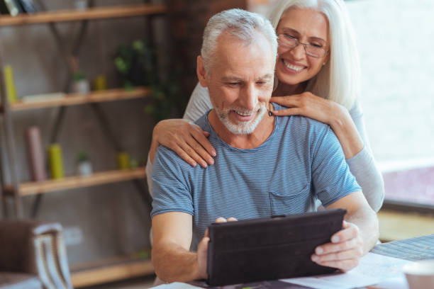 Retired positive man using tablet with his wife Happy family days. Waist up of a cheerful retired man using tablet and sitting at the table while resting together with his loving wife at home baby boomers stock pictures, royalty-free photos & images