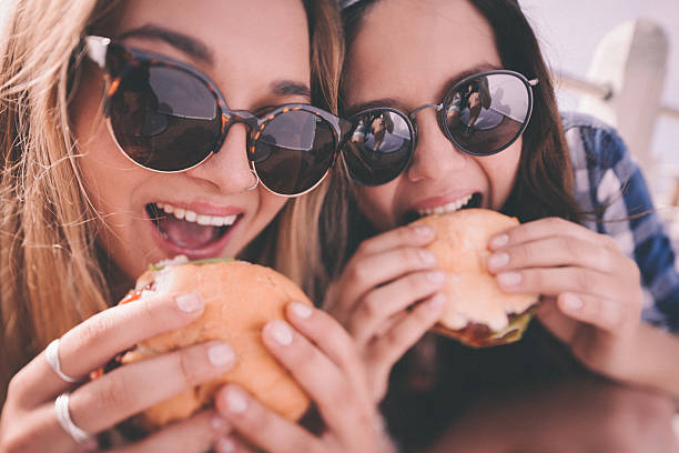 Retro style shot of teenage girl best friends eating burgers Closeup portrait of two teenage girls who are best friends, wearing hipster sunglasses about to take bites out of large burgers outdoors with a retro style develop biting photos stock pictures, royalty-free photos & images
