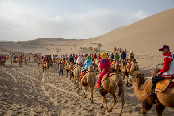 Riders in crowds at camel rides, Singing Sand Mountain, Taklamakan Desert, Dunhuang, China Dunhuang, Gansu, China - September 12, 2018 : Camel riders on dunes  at Singing Sand Mountain dunes of Taklamakan Desert, Dunhuang, China. silk road stock pictures, royalty-free photos & images