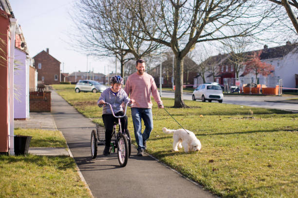 Riding his Bike A boy with down syndrome rides his bike through a residential district whilst his father walks beside him with the dog. adult tricycle stock pictures, royalty-free photos & images