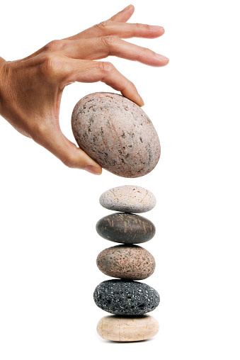 Concept of a risky proposition, reckless move, danger, poor judgement, or multi-tasking miscalculation represented by human hand placing a large rock on a precariously balanced stack of small stones. Positioning requires care but may be careless, is piling opportunity to  outweigh fragility, and arranging opportunity over ignorance or failure.