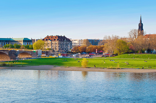 Riverbank of Elbe river in Dresden, Germany. Many people sitting on fresh green lawn and enjoying looking at the shore. Cloudless spring day. Dome of Martin Luther Kirche. Shot at golden hour