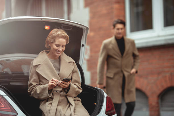 Road trip Handsome couple is getting ready for a road trip. The woman is sitting on the edge of the car trunk and using smartphone and the man is coming from the distance with the suitcase. georgijevic frankfurt stock pictures, royalty-free photos & images
