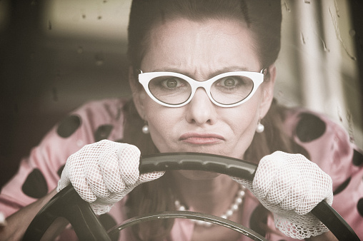 Mature woman who wearing white retro styled glasses, lace gloves, pearl necklace and doted blouse driving an oldtimer car in the rain - 1950 style. Horizontal sepia toned image.