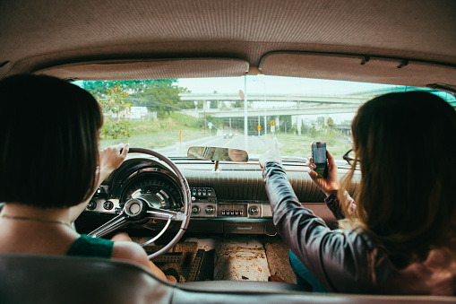 Lifestyle stock image of two young woman driving vintage american car. The passenger uses smart phone to pull up directions on the GPS app. 