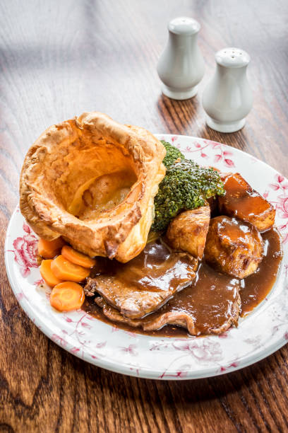 roast beef complete Sunday roast platter with sliced roast beef, carrots, broccoli, potatoes, yorkshire pudding and a mountain of gravy in a white plate on a wooden background with salt and pepper in the background roast dinner stock pictures, royalty-free photos & images