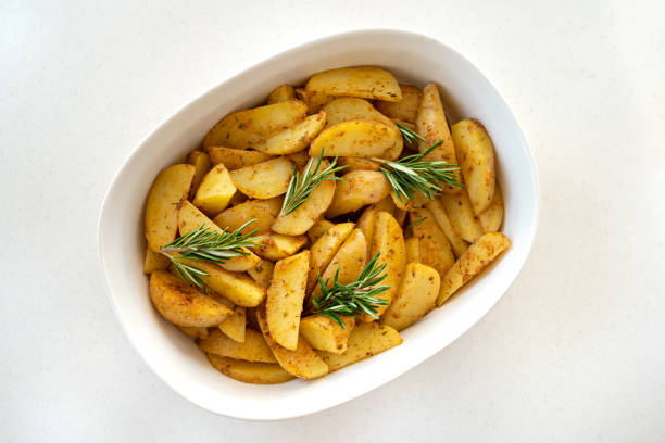 Roasted potato wedges with spicy herbs, spices and pieces of fresh rosemary on the white porcelain baking dish stock photo